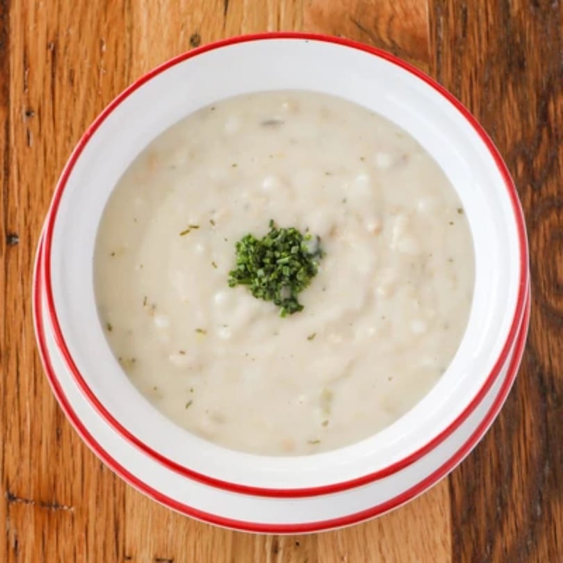 Creamy white soup with green herbs in a white bowl with red trim on a wooden table.