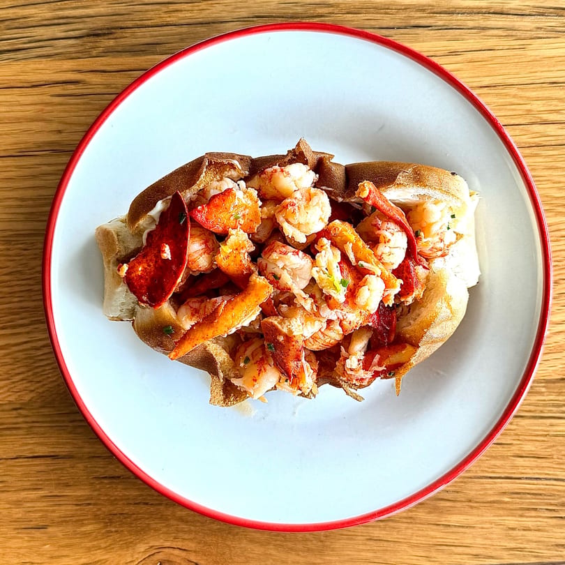 A lobster roll with generous chunks of lobster meat and claws on a white plate with a red rim.
