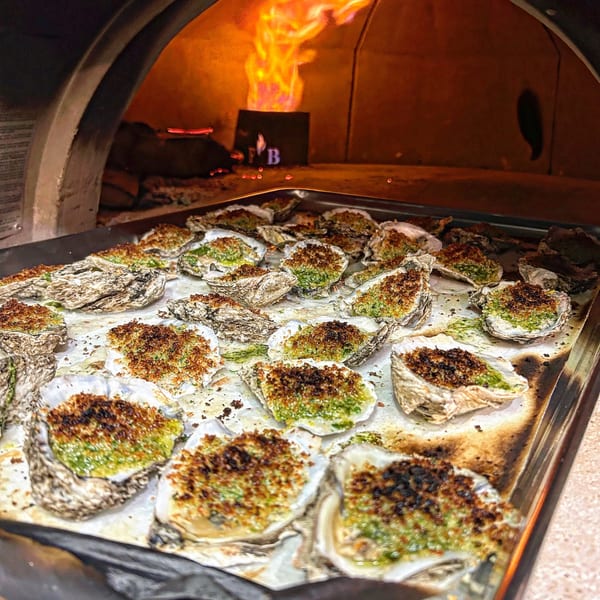 Baked oysters with green topping and breadcrumbs on a tray in front of a wood-fired oven.