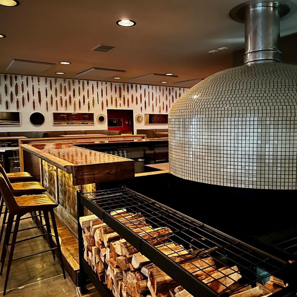 A tiled pizza oven, stacked firewood, and a bar with stools in the GOOMAH restaurant.
