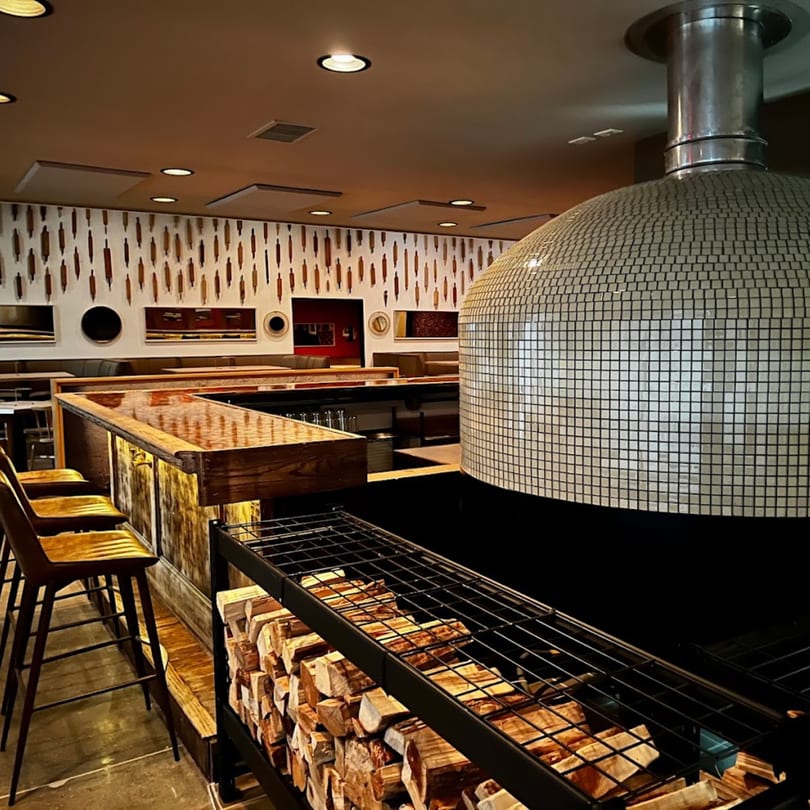 A tiled pizza oven, stacked firewood, and a bar with stools in the GOOMAH restaurant.